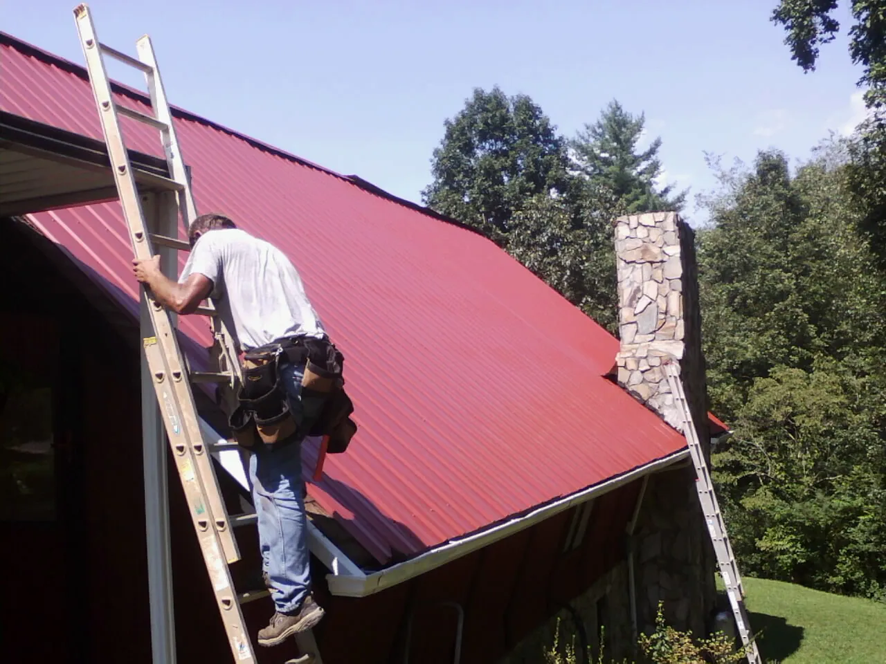 Roofer on a ladder beside a red metal roof and stone chimney — Soffit & Fascia Repair by Roofing Brothers in Templeton